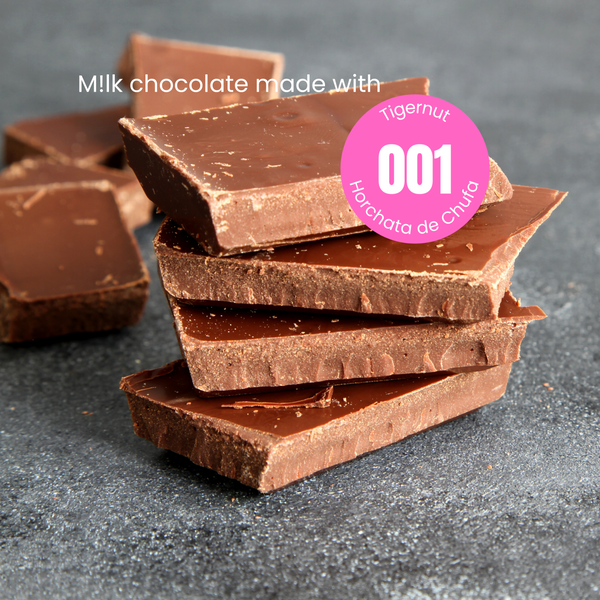 4 rectangular pieces of plant-based milk chocolate stacked on a grey stone table with some more pieces scattered in the background. The words 'milk chocolate made with’' in white text over the chocolate. The word milk is written with an exclamation mark indicating plant-based. Overlapping the chocolate is a pink circular sticker label overlay with text in white with 001 in the centre with the words Tigernut indicating the recipe of the powder that was used to make this chocolate.