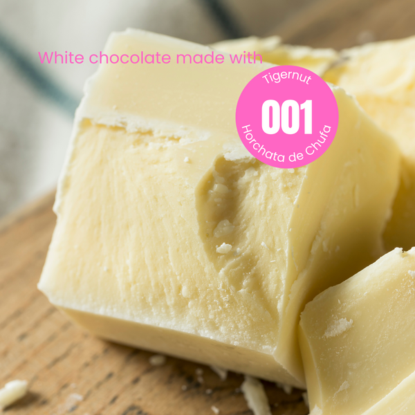 Large chunks of white chocolate on a wooden table. The words 'White chocolate made with' in pink are written over the chocolate. Overlapping the chocolate is a pink circular sticker label overlay with number 001 in the centre with the words Tigernut indicating the recipe of the powder that was used to make this chocolate.