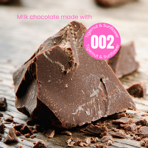 A pyramid shaped chunk of rough cut plant-based milk chocolate with smaller crumbled bits scattered on a wooden table. The words 'milk chocolate made with' in white written above the chocolate.  The word milk is written with an exclamation mark indicating plant-based. Overlapping the chocolate is a pink circular sticker label overlay with number 002 in the centre with the words Tigernut and Sunflower indicating the recipe of the powder that was used to make this chocolate.