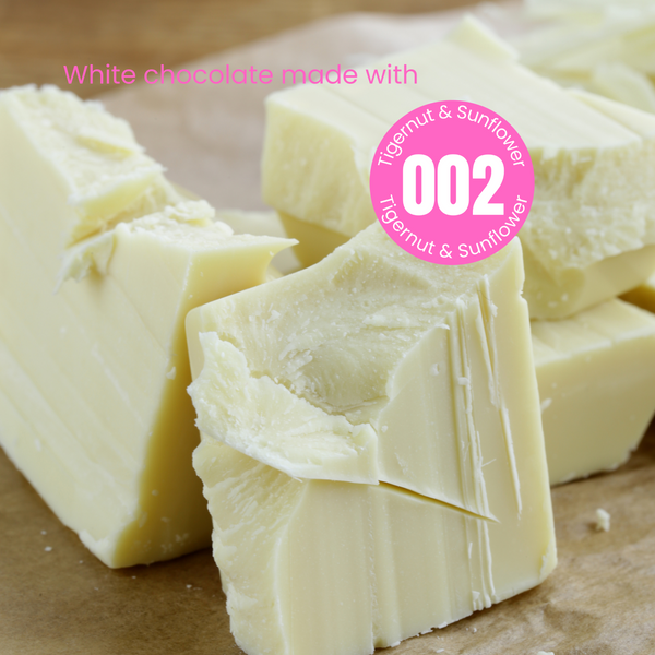 Rough chunks of plant-based white chocolate on a wooden table. The words 'White chocolate made with' in pink are written over the chocolate.  Overlapping the chocolate is a pink circular sticker label overlay with number 002 in the centre with the words Tigernut and Sunflower indicating the recipe of the powder that was used to make this chocolate.