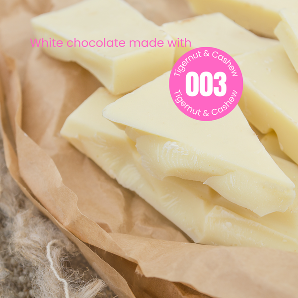 Rough broken pieces of plant-based white chocolate on brown paper. The words 'White chocolate made with' in pink are written over the chocolate.  Overlapping the chocolate is a pink circular sticker label overlay with number 003 in the centre with the words Tigernut and Cashew indicating the recipe of the powder that was used to make this chocolate.